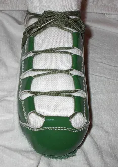 A person wearing Celtic Choice Patent Leather Green and white shoes on a bed.